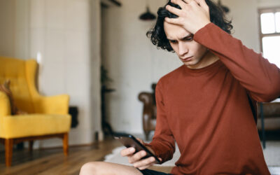 Frustrated young man having stressed facial expression, keeping hand on his head, holding cell phone, reading bad news or receiving spam message. Negative human emotions and electronic gadgets